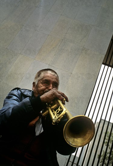 MEXICO, Mexico City, "In downtown Mexico City on the side of one of the main Plazas namely the Zocolo sits an old musician. He is purched on a small wooden stool almost propped up against a grey stone wall. Next to the wall in the background stands a metal grill protecting a kept private garden. The musician is an ageing local dressed in a blue shiny puffer jacket, jeans and an off-white shirt. His hair has been cut close and is grey at the front and sides. As he holds the fairly worn trumpet up to his lips his cheeks inflate and the wrinkles and expression lines become apparent in his dark face. His large yet agile fingers lie over the buttons of the trumpet tinkling away. As locals pass by they hear the local classics that this Mexican is playing."