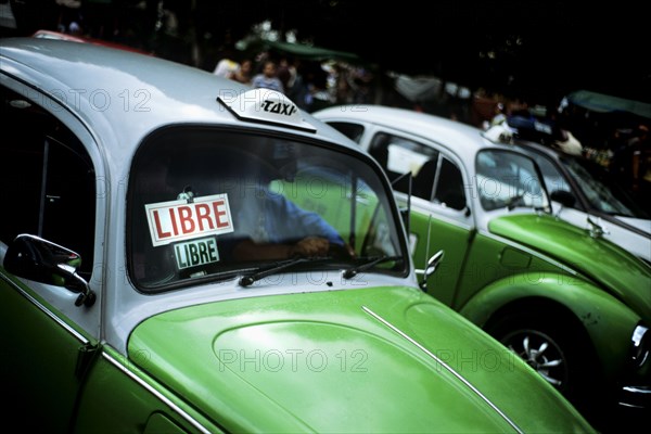 MEXICO, Mexico City, Down town Mexico City on the north side of the Zocolo sits a line of the world famous green Beatle taxis. All taxis in Mexico City are of this design and being the middle of the day business seems to be a little slow. In the background one can make out the blurs of a bustling City Centre whereas in the foreground are quietly parked a whole line of taxis. Each one almost identical. Painted halfway in traditional Mexican green with their top halves in white. On top of each roof sits a 'taxi' light and in each windscreen a stuck on sign saying 'libre' or 'available'. Although the taxis will more than likely be fairly old their wind mirrors and alloy wheels shine in the afternoon light as the actual insides of the cars are in shadow.