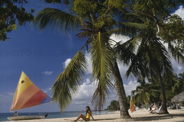 DOMINICAN REPUBLIC, Casa de Campo, Las Minitas Beach lined with palm trees and windsurfs with a woman sat on the sand under a palm tree in the foreground