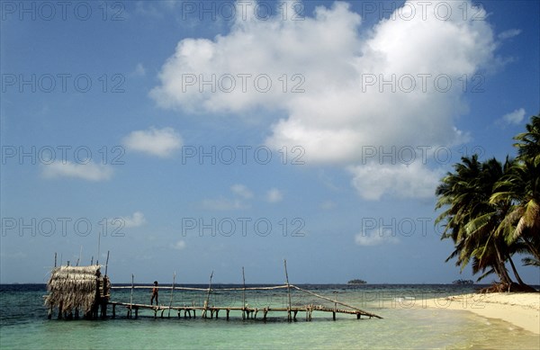 PANAMA, San Blas Islands, Isla Pequena, "The 'Comarca de Kuna Yala' is a narrow 226km strip of the Caribbean coast of Panama that includes the Archipelago de San Blas. The area consists of around 400 islands and are home to the Kuna, who run San Blas as a 'comarca' - an autonomous region with minimal interference from the Panamanian government on the mainland. They have their own system of governance, philosophies and decision-making, whilst maintaining their own economic systems, language, customs and cultures. Originally from Colombia, they fled to the islands when the Spanish first arrived in the 16th Century. In this particular shot we see rich blue skies with sporadic sprays of fluffy clouds and paradise like turquoise clear waters. The sand is soft, clean  and white and the whole of the island is covered in 'cocoteros' or coconut palmes of all different shapes and sizes. In the middle of shot almost attached to the island sits a wooden pier or jetty. It sits on a collection of st...