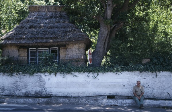 PORTUGAL, Madeira, Camacha, Traditional thatched cottage beside the Largo do Achada the central square with a man sat on the pavement next to a white wall