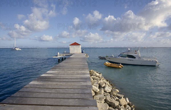 GUADELOUPE, St Froulon, Wooden jetty stretching out to sea with a man sat next to a hut at the end of the jetty with boats moored on the water nearby