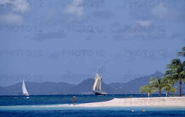 GRENADINES, Petit St Vincent, Palm Island Resort. Man walking on sandy outstretch of beach surrounded by turquoise sea with boats on water