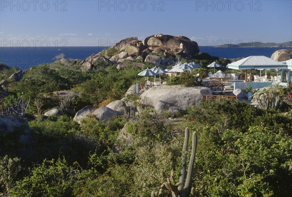 BRITISH VIRGIN ISLANDS, Virgin Gorda, The Baths. Granite boulders amongst plants and vegetation with chairs and sun shades from a restaurants open air verandah in the middle. The ocean seen in the distance