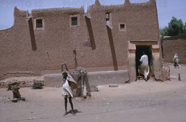 NIGERIA, Kano, Traditional mud Hausa dwelling with child standing with up-stretched arms outside and man entering through open doorway.