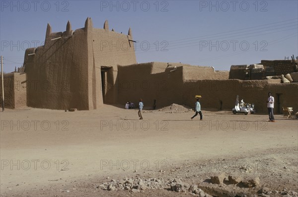 NIGERIA, Kano, Ancient mudbrick city walls and passing people and street trader carrying tray of fruit on his head.