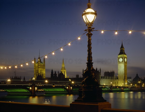 ENGLAND, London, Westminster. Houses of Parliament and Westminster Bridge illuminated at night seen from across the Thames with riverside lighting in the foreground.