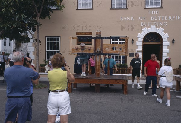BERMUDA, St George, Tourists in kings Square with people in the foreground taking pictures of a man standing in a wooden execution guillotine outside The Bank of Butterfield Building