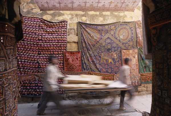 INDIA, Rajasthan, Jaisalmer, Jaisalmer Fort. Two men pushing a cart through a narrow street with textile rugs hanging from the walls