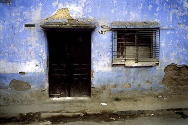 GUATEMALA, Quetzatenango, Xela, "A backstreet in the outskitrs of the Guatemalan town of Xela. A characteristic but crumbling house sits baking in the sun, infront lies a dishevelled pavement and a dry dusty road. The house itself is worn but glows a beautiful blue colour in the afternoon sun. On the front wall of the house sits the main entrance, a comparatively grand black double wooden door. On the floor of the entrance are a few old but relatively shiny ceramic tiles. To the right sits the number of the house and then a double window behind the standard Latin-American metal bars which cover all houses whuch face a public street. The metal grill is black and attached firmly to the stonework. A lot of the blue paintwork has flaked off and revelas the pld stonework beneath. On the other side of the door is a small metal plaque which show the name of the owner of the house, again, a very Latin characteristic."