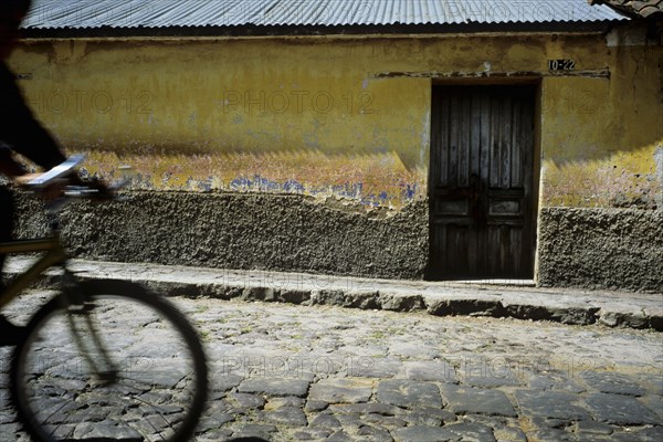 GUATEMALA, Quetzatenango, Xela, "A backstreet in the outskitrs of the Guatemalan town of Xela. A characteristic but crumbling house sits baking in the sun, infront lies a dishevelled pavement and a dry dusty cobbled road. The house itself is worn but glows a beautiful yellow colour in the afternoon sun. Beneath the flaking paint one can see the years of history that the building carries. Orange and blue faded paintwork can all be seen. On the front wall of the house sits the main entrance, a small wooden door. The roof of the house is made of corrugated iron panels, again very characteristic if this area of Latin America. At the front of shot passes a local indigenous young man on his bicycle. The handlebars shine in the sun whilst his body and face remain in silohette."