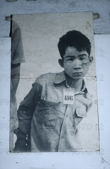 CAMBODIA, Phnom Penh, Tuol Sleng Museum. Photograph of a young male prisoner in a Khmer Rouge prison wearing a number tag around his neck