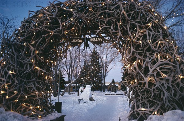 USA, Wyoming, Jackson Town Square. View through Elk Horn Archway decorated with lights towards snow and a Christmas tree.