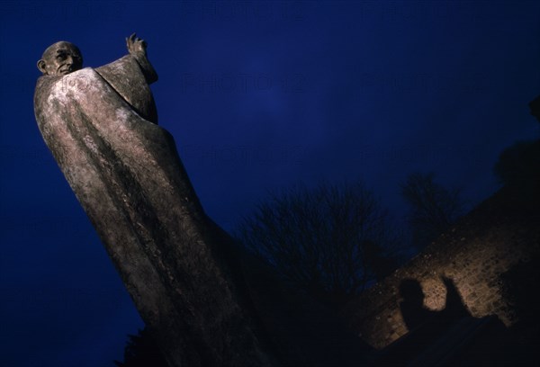 ENGLAND, West Sussex, Chichester, Statue of Saint Richard partially illuminated at night in Cathedral grounds with shadow reflecting on nearby wall.