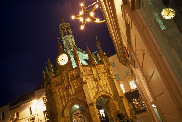 ENGLAND, West Sussex, Chichester, Angled view of The Cross at night  with star shaped Christmas light attached to nearby building with clock reflected in glass window.