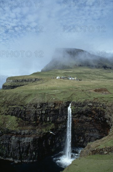 DENMARK, Faeroe Islands, River Gazadalla, Waterfall and rocky coastline with houses built on green landscape
