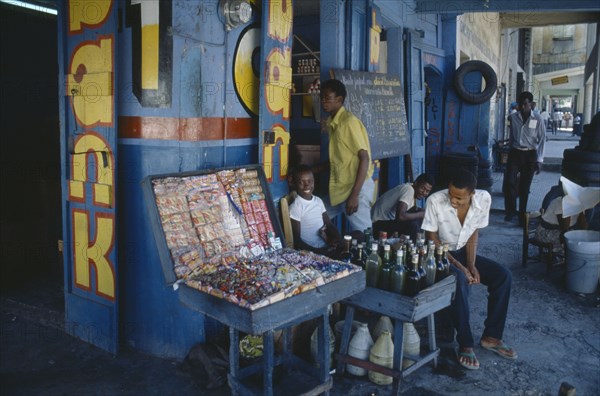 HAITI, Port au Prince, Children and young men outside shop of sweets and drinks vendor with blue and yellow painted exterior.