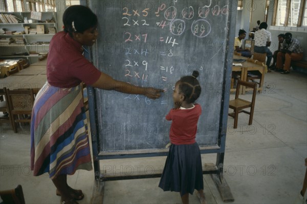 GRENADA, Education, Female teacher pointing out 2 x table written in coloured chalk on blackboard to little girl.