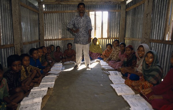 BANGLADESH, Char Kukri Mukri, Teacher and pupils in village school.  Students sit on earth floor of corrugated metal hut.