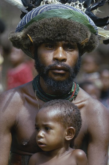 PAPUA NEW GUINEA, New Guinea, "Melpa tribesman wearing hat decorated with fur, feathers and leaves with young child sitting in front of him."