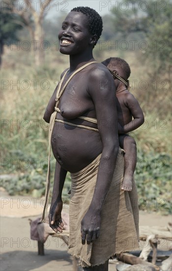 ETHIOPIA, West, Gambella, Uduk refugees from Sudan.  Smiling pregnant woman carrying baby on her back in cloth and wood sling.  Note scarification on stomach.