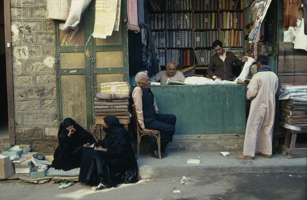 EGYPT, Market, "Market scene.  Vendors, customer and women in conversation outside pavement stall selling fabric."