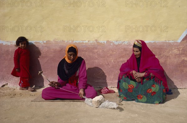 EGYPT, Western Desert, Bedouin, "Women and child in colourful dress spinning wool, sitting against faded yellow and ochre coloured wall of building."