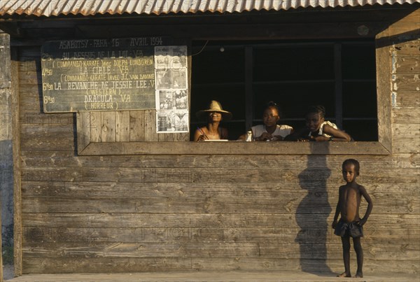 MADAGASCAR, Fort Dauphin, Child standing outside local cinema ticket office with three girls at open window and blackboard on outside wall with film listings in French.