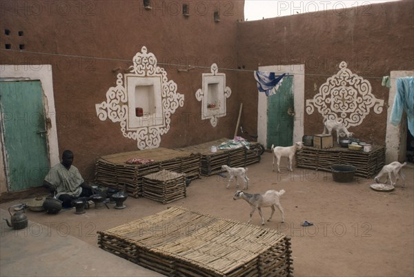MAURITANIA, Oualata, "Man preparing food in courtyard of buildings with traditional bas-relief ornamentation of applied gypsum, white and red clay with turquoise painted doors, washing line and goats."