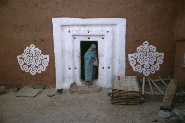 MAURITANIA, Oualata, "Woman stepping out from doorway with white surround and traditional bas relief motifs on each side of applied gypsum, white and red clay."