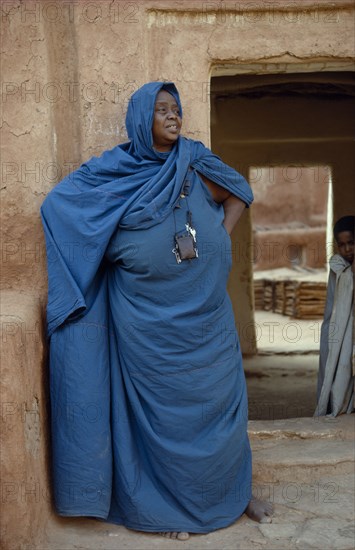 MAURITANIA, Oualata, Portrait of woman wearing turquoise robe and head covering standing in front of mud brick building with child framed in doorway behind her.