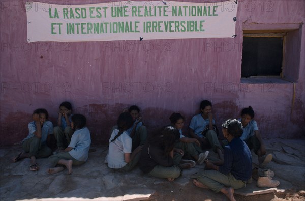 WESTERN SAHARA, SADR, Sahrawi schoolgirls sitting against pink plastered wall under banner with slogan in French: RASD is a national and international irreversible reality.