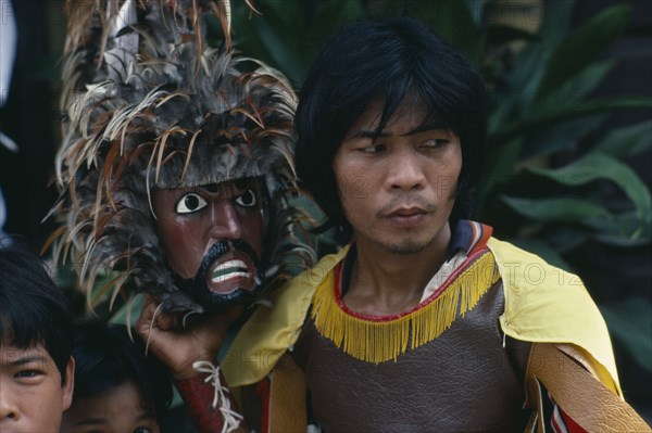 PHILIPPINES, Marinduque, Boac, "Man taking part in re-enactment of the crucifixtion during Easter week, head and shoulders portrait holding mask with feather head-dress."