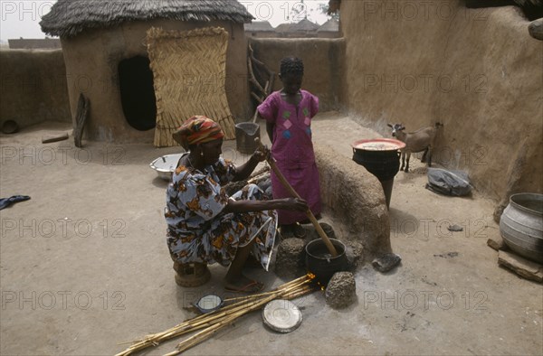 GHANA, Chereponi, "Woman cooking over open stove watched by young girl in small, walled yard with goat kid and thatched mud brick hut behind."