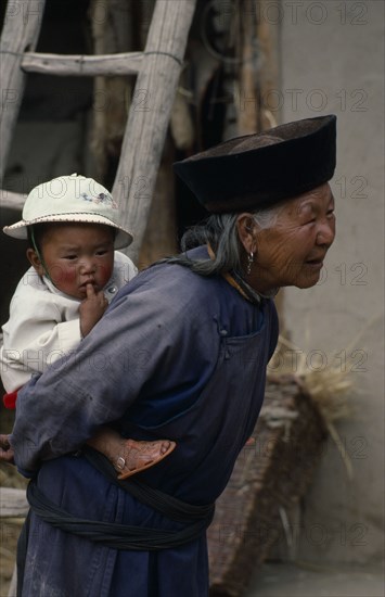 CHINA, Qinghai Province, Huzhu District, Elderly Tu nationality Lamaist woman of Yellow Hat Buddist sect carrying child on her back.