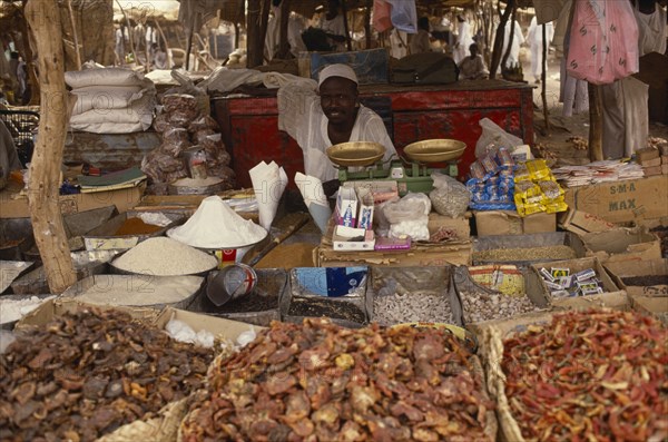 SUDAN, North Darfur, El Fasher, Market trader sitting behind scales and display of dried foodstuffs and other goods.