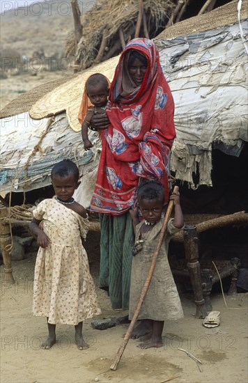 SUDAN, North East, Gadem Gafriet Camp, Beni Amer nomad refugee woman and children outside tent in desert camp with circular prayer mat on roof made from woven palm leaves or Birrish.