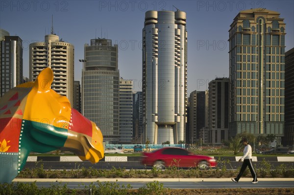 UAE, Abu Dhabi, "City centre skyline along the Corniche. Statue of a colourful patchwork camel, red car on road and a pedestrian."