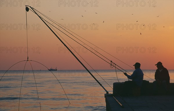 ITALY, Tuscany, Castiglione della Pescaia, Fishermen on harbour wall at sunset with distant fishing boat.