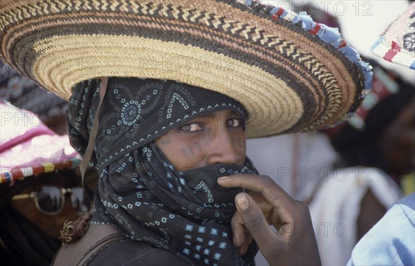 20075137 NIGERIA People Men Portrait of Wodaabe man wearing wide brimmed hat and scarf over his head and mouth with hand raised to face.