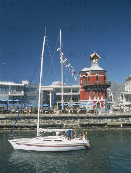SOUTH AFRICA, Western Cape, Cape Town, Victoria and Alfred Waterfront. View across water with a yacht sailing past towards a Victorian Gothic style Clock Tower with people sat under parasols along the waterside.