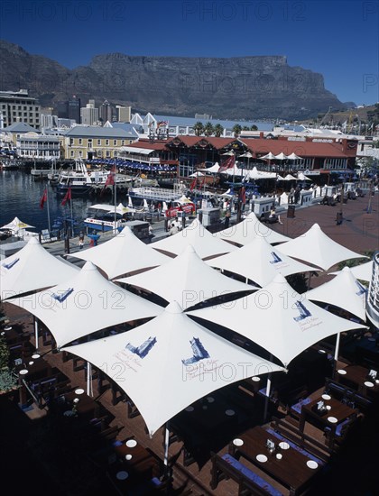 SOUTH AFRICA, Western Cape, Cape Town, Victoria and Alfred Waterfront with view across white  awnings towards waterside restaurant and bars with boats docked in marina. Table Mountain behind
