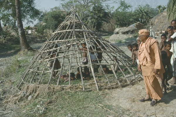 INDIA, West Bengal, Local guru or holy man on village tour inspecting frame of hut waiting to be thatched.