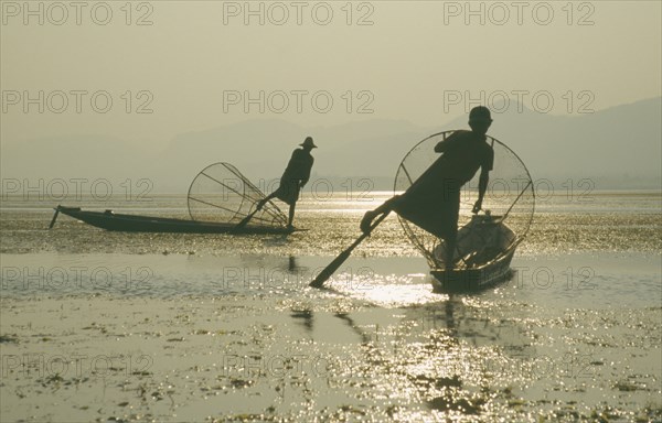 MYANMAR, Shan State, Inle Lake, Silhouettes of two Intha fishermen with Sun’s reflection on water.