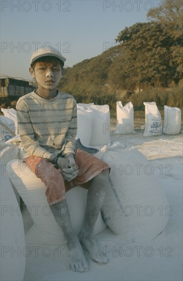BURMA, Mandalay , "Portrait of a young boy worker at Mandalay Dock sat on bags of cement covered in dust, wearing a baseball cap. Banks of the Irrawaddy"