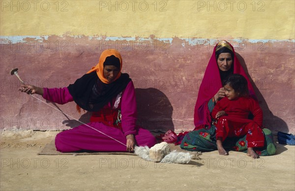 EGYPT, Western Desert, Settled Bedouin woman spinning wool with older woman and child sitting beside her against faded ochre and terracotta painted wall.