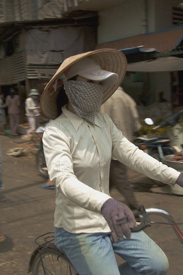 VIETNAM, South, Ho Chi Minh City, Woman wearing a conical hat with a scarfe covering her mouth riding a bycicle