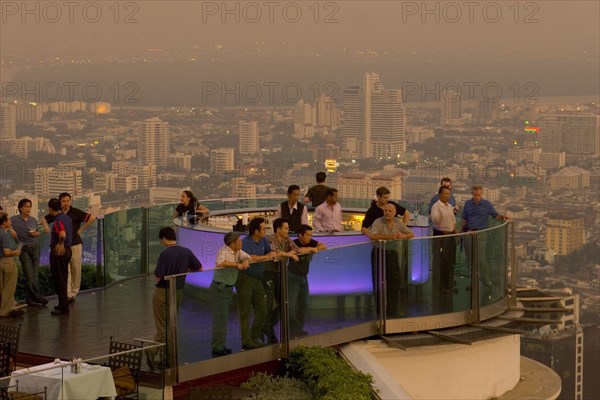 THAILAND, Bangkok, Sorocco Restaurant. People standing next to a lit up bar and looking over balcony with the Bangkok Skyline in the background.