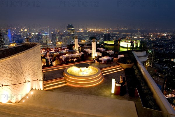 THAILAND, Bangkok, Sorocco Restaurant. View down steps towards people dining at tables lit up with light displays and the Bangkok Skyline in the background.