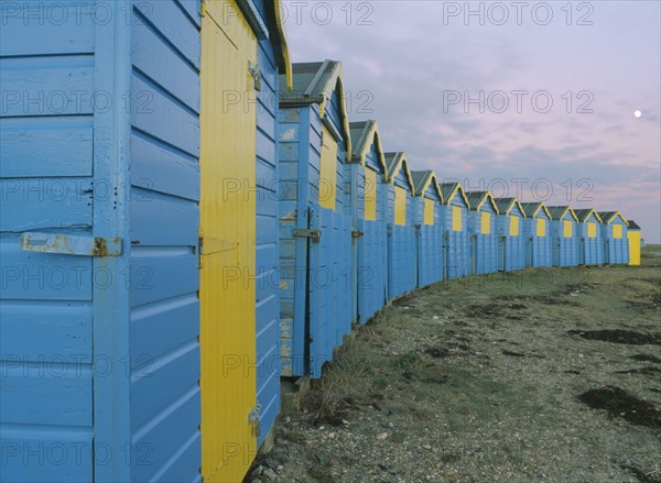 ENGLAND, West Sussex, Littlehampton, Crescent of blue and yellow beach huts in evening light with moon showing.